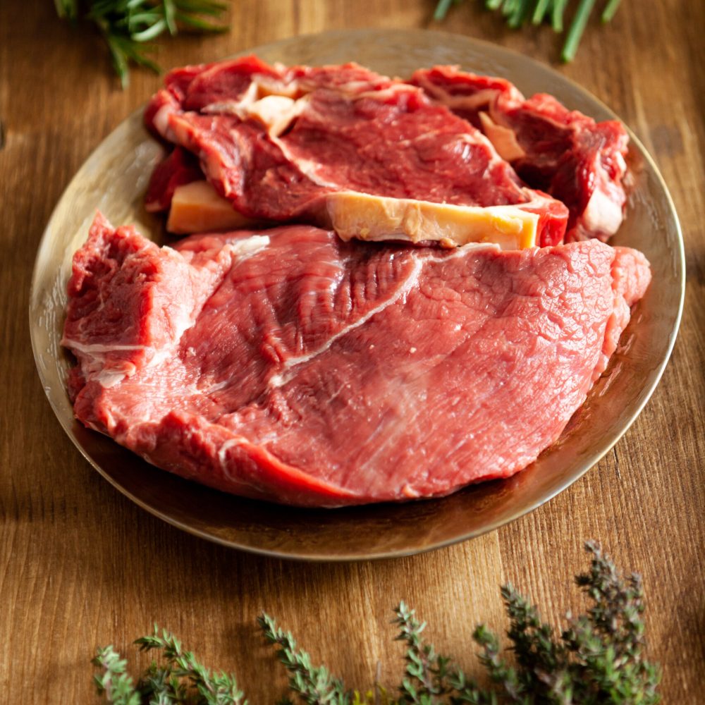 Different types of red meat in a vintage plate next to green vegetables and hersb on wooden table. Dinner preparation.