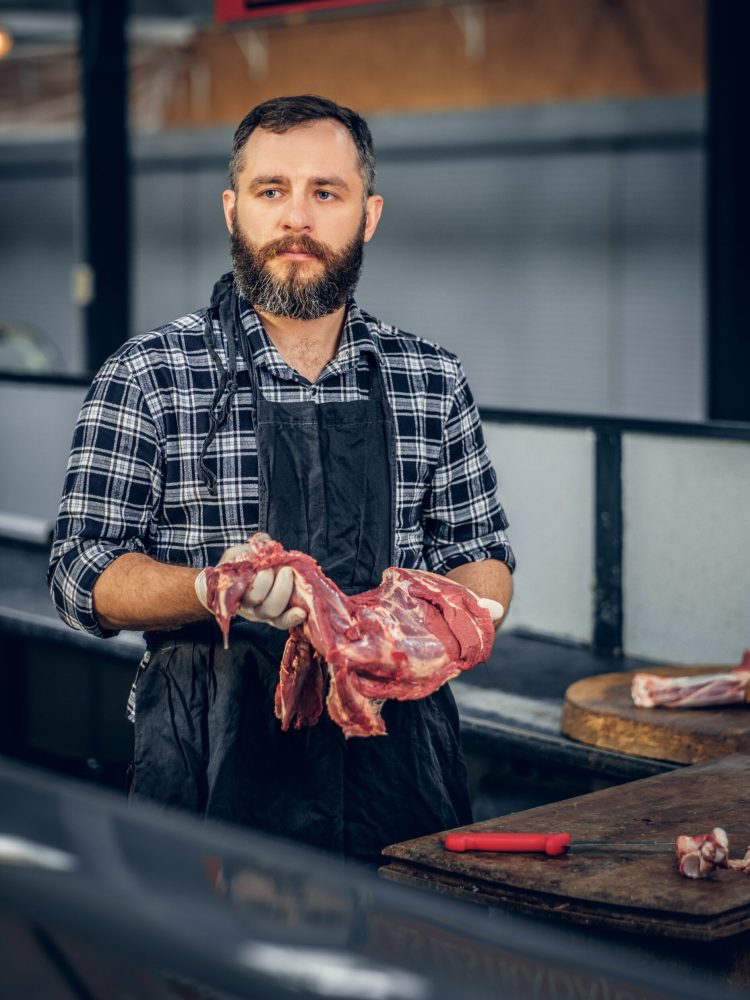 Portrait of a bearded meat man dressed in a fleece shirt holds fresh cut meat in a market.