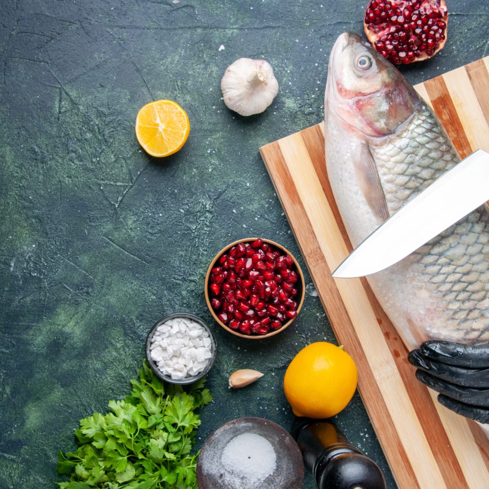 top-view-chef-with-black-gloves-cutting-raw-fish-cutting-board-pepper-grinder-pomegranate-seeds-bowl-kitchen-table-copy-place